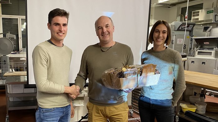 Bezirksversammlung des lvh-Bezirks Brixen Drei Personen, zwei Männer schütteln Hände, eine Frau hält Geschenkkorb in einem Raum