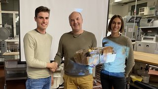 News Drei Personen, zwei Männer schütteln Hände, eine Frau hält Geschenkkorb in einem Raum