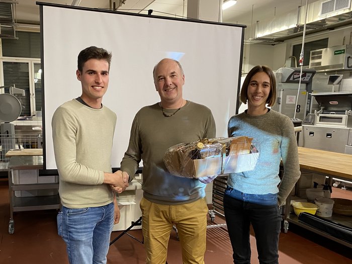 Riunione del circondario Bressanone di lvh.apa Tre persone, due uomini si stringono la mano, donna tiene un cesto regalo in una stanza