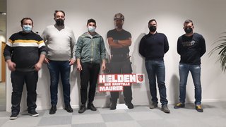 News Fünf Männer mit Masken stehen neben einer Bauarbeiter-Pappfigur mit Schild Helden der Baustelle Maurer