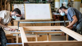 News Zwei Handwerker mit Masken arbeiten an einem Holzrahmen in einer Werkstatt.
