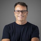 Stefano Mason Uomo sorridente con occhiali e maglietta blu su sfondo grigio