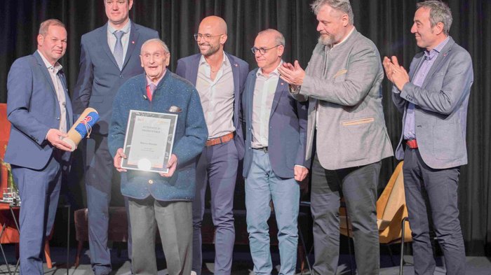 “Artigiani. Mani e Cuore” Uomo anziano riceve un premio con certificato circondato da uomini che applaudono