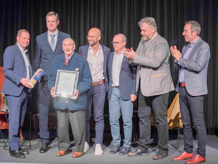 “Artigiani. Mani e Cuore” Uomo anziano riceve un premio con certificato circondato da uomini che applaudono