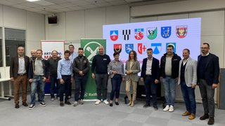 News Gruppe von Menschen steht in einem Raum vor Bannern und einem Bildschirm mit Wappen