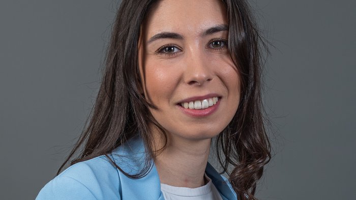 3670 neue junge Fachkräfte Porträt einer lächelnden Frau mit braunen Haaren und blauem Blazer vor grauem Hintergrund