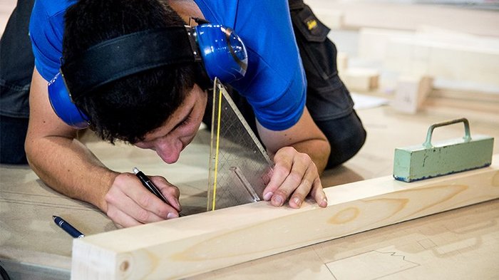Holzbaumeister und Zimmerer im lvh: „Die Ausbildung ist der Schlüssel“ Mann trägt Gehörschutz und zeichnet mit Geodreieck auf Holzstück