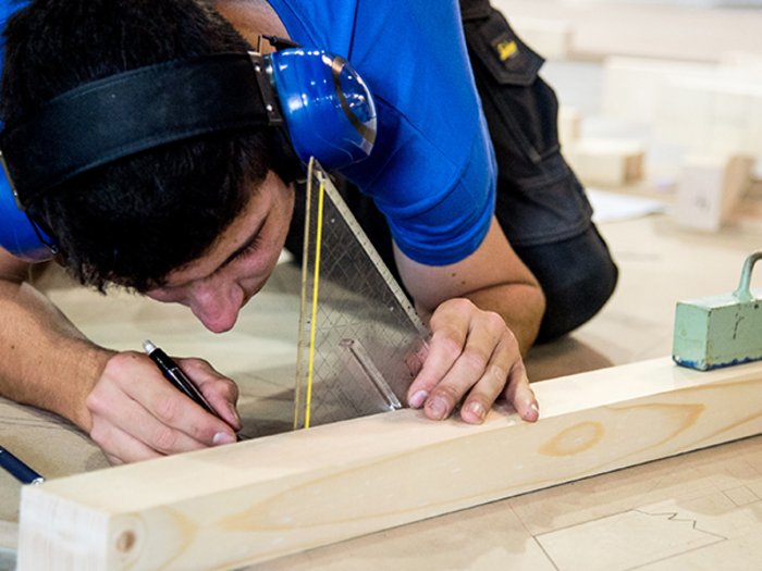 Holzbaumeister und Zimmerer im lvh: „Die Ausbildung ist der Schlüssel“ Mann trägt Gehörschutz und zeichnet mit Geodreieck auf Holzstück