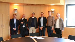 News Sechs Männer in Geschäftskleidung stehen in einem Besprechungsraum vor einem runden Tisch