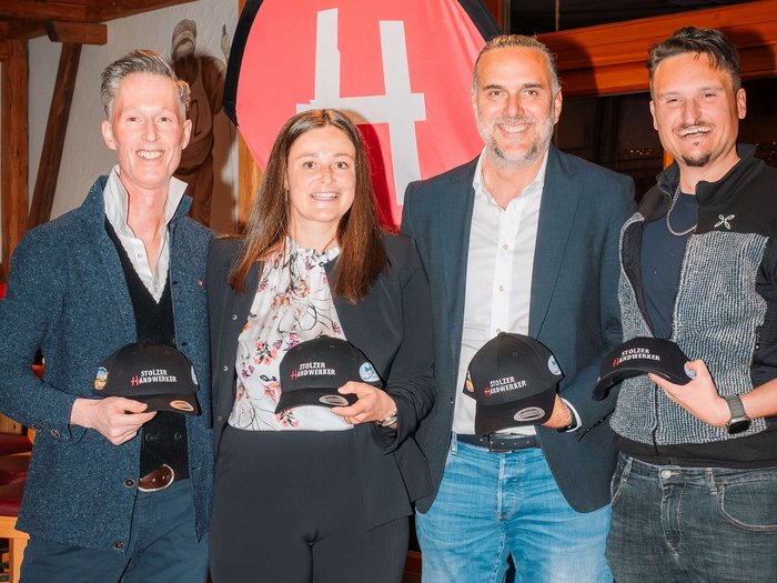Erfolgreiche lvh-Ortsversammlung in Toblach: Ein Jahr voller Engagement und Zusammenarbeit Vier Personen halten Stolzer Handwerker Mützen vor einem roten Banner