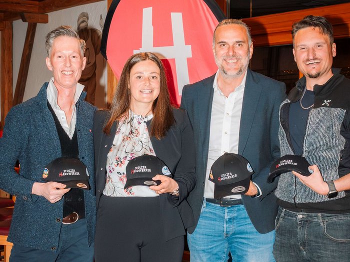 Successo della riunione comunale del gruppo lvh.apa a Dobbiaco: un anno ricco di impegno e collaborazione Quattro persone tengono cappellini Stolzer Handwerker davanti a uno striscione rosso