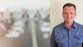 News Porträt eines lächelnden Mannes in blauem Hemd vor unscharfem Firmenmeeting