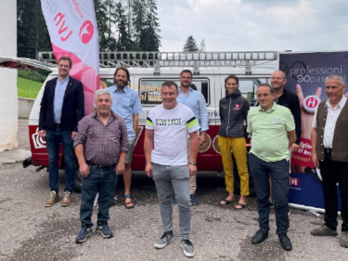 Junges Südtiroler Handwerk - von lokal zu international Gruppenfoto von neun Personen vor Fahnen und einem Anhänger im Freien