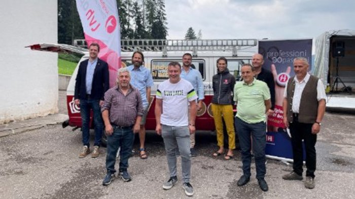 Junges Südtiroler Handwerk - von lokal zu international Gruppenfoto von neun Personen vor Fahnen und einem Anhänger im Freien