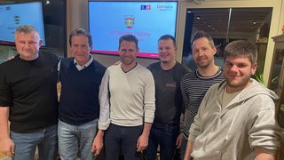 News Sechs Männer stehen lächelnd in Innenraum vor Bildschirm mit Schrift Ortsgruppe Truden