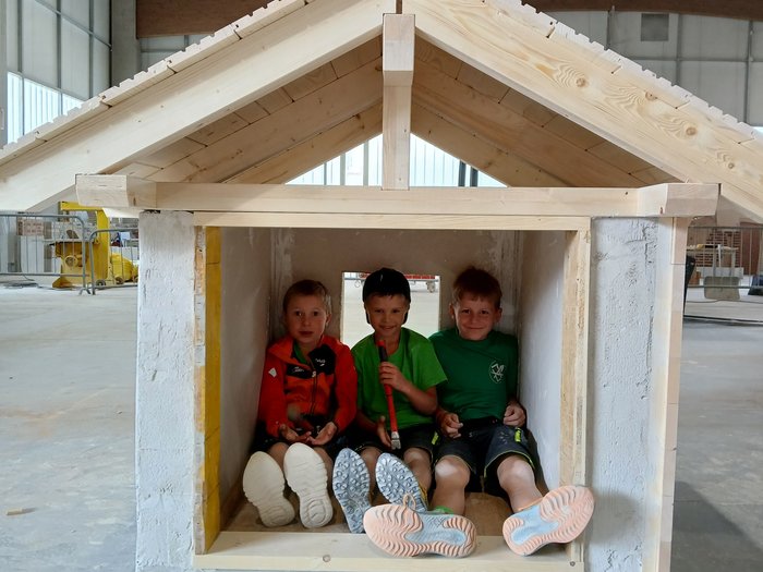 Mini-Baustelle, große Meisterwerke – KidsAcademy mauert in Bruneck Drei Kinder sitzen in einem kleinen Holzhaus in einer Halle