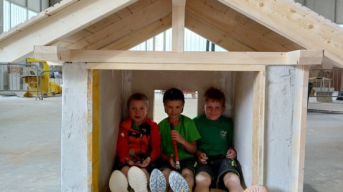 Mini-Baustelle, große Meisterwerke – KidsAcademy mauert in Bruneck Drei Kinder sitzen in einem kleinen Holzhaus in einer Halle