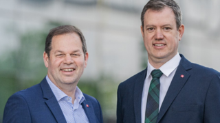 News Zwei Männer in Anzügen lächelnd vor einem Gebäude