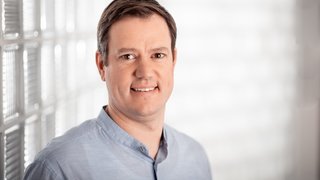 News Porträt eines lächelnden Mannes in blauem Hemd vor hellem Hintergrund