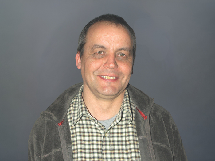 Holzbaumeister/innen und Zimmerer/innen begrüßen Förderung für privaten Holzbau Mittelalter Mann mit kariertem Hemd und Jacke vor grauem Hintergrund lächelnd