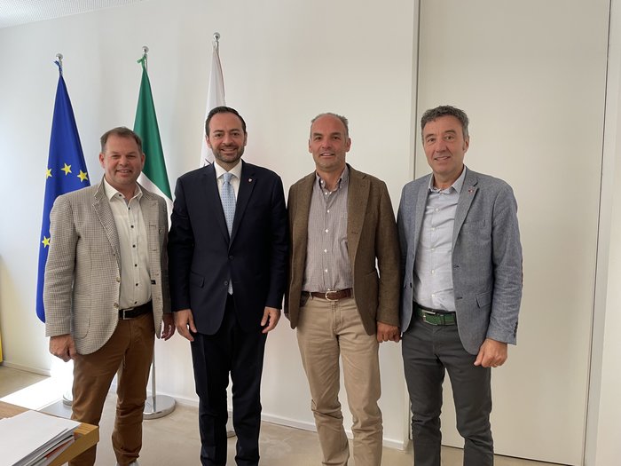 Südtiroler Warentransport: Branche fordert Bürokratieabbau und bessere Ausbildungsbedingungen Vier Männer in Anzügen vor EU-, Italien- und weiteren Flaggen