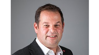 News Portrait eines lächelnden Mannes in weißem Hemd und dunklem Sakko vor grauem Hintergrund