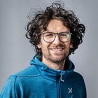 Orthopädieschuhmacher Porträt eines lächelnden Mannes mit lockigem Haar und Brille in blauem Hoodie