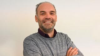 News Uomo sorridente con maglione grigio e braccia incrociate davanti a sfondo bianco