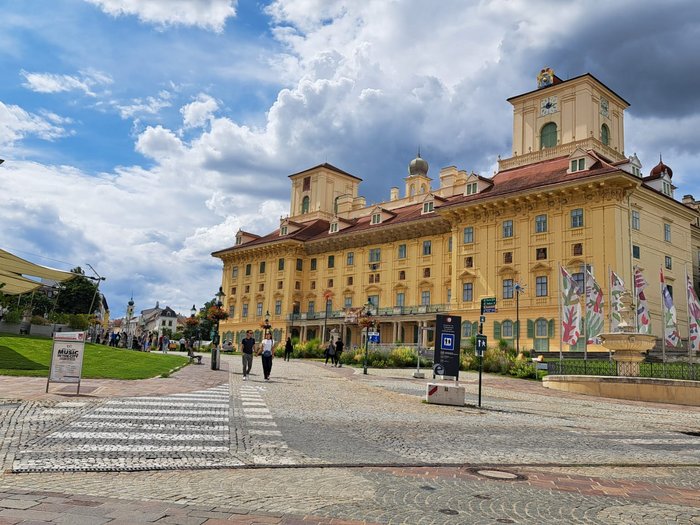 Viaggio culturale nel Burgenland – Opera e musical come fulcro del programma Edificio storico giallo con cielo nuvoloso, marciapiede e bandiere