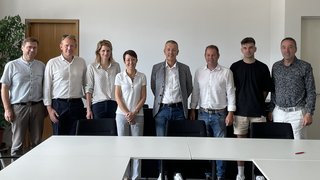 News Neun Personen in einem Besprechungsraum stehen hinter weißen Tischen