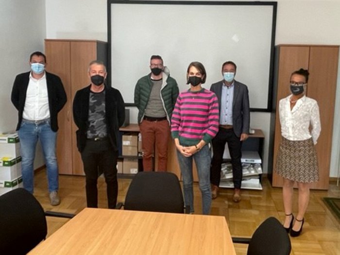 Über öffentliche Aufträge in Brixen Sechs Personen mit Masken in einem Besprechungsraum stehen vor Leinwand und Schränken