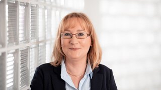 News Frau mit Brille und Blazer vor hellem Hintergrund lächelnd