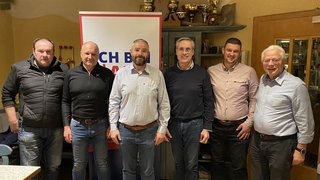 News Sechs Männer stehen vor einem Banner und Regalen mit Pokalen in einem Raum