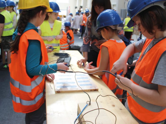 Erlebniswelt Baustelle Kinder in Schutzhelmen und Warnwesten basteln mit Draht an einem Holztisch
