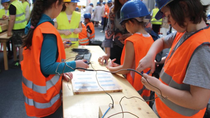 Erlebniswelt Baustelle Kinder in Schutzhelmen und Warnwesten basteln mit Draht an einem Holztisch