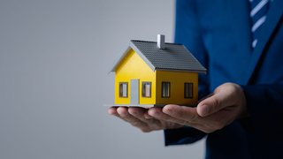 News Person hält ein Modellhaus in den Händen vor neutralem Hintergrund