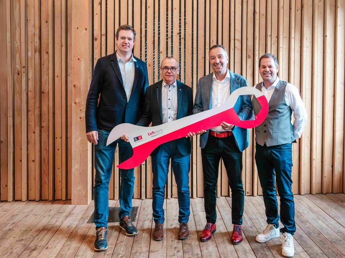 Nominato il nuovo direttore di lvh.apa: Walter Pöhl assume la direzione Quattro uomini in abiti formali tengono una grande chiave inglese rossa davanti a una parete di legno