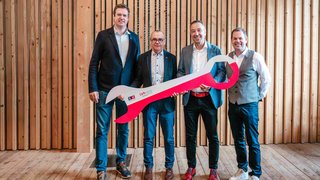 News Vier Männer in formeller Kleidung halten einen übergroßen roten Schraubenschlüssel vor Holzwand