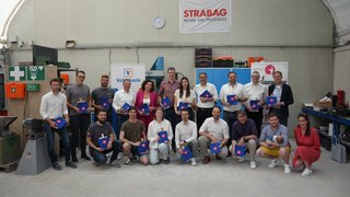 News Gruppenfoto von 19 Personen mit blauen Taschen in einer Werkstatt