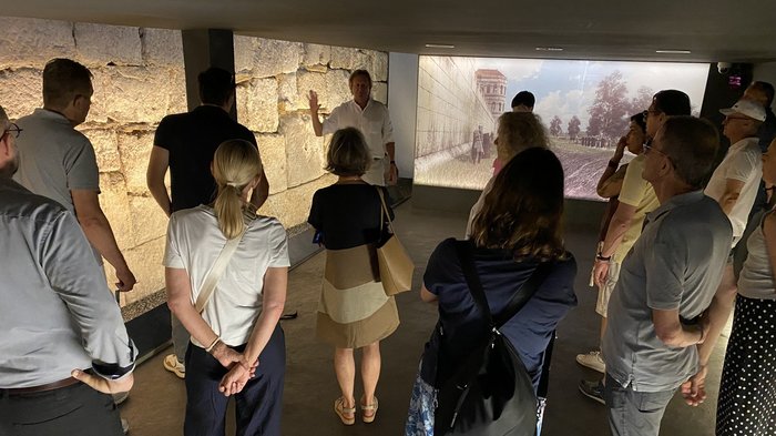 Cinque Paesi, un obiettivo: i tecnici termosanitari e della ventilazione puntano sulla cooperazione europea Gruppo ascolta una guida davanti a muro antico e proiezione su parete