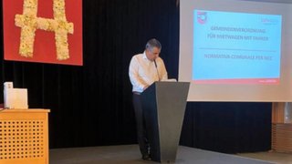 News Mann hält Vortrag auf Bühne neben Präsentationsfolie mit kommunaler Verordnung