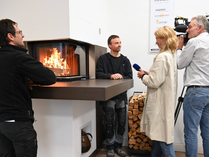 Heizen mit Holz: Landesinitiative gemeinsam mit Kaminkehrer/innen im lvh wird fortgesetzt Interview vor einem Kamin mit brennendem Holz und Holzstapel im Raum