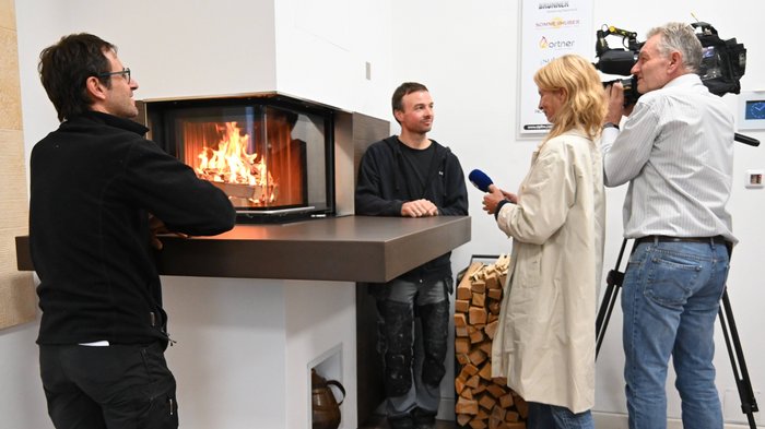 Heizen mit Holz: Landesinitiative gemeinsam mit Kaminkehrer/innen im lvh wird fortgesetzt Interview vor einem Kamin mit brennendem Holz und Holzstapel im Raum