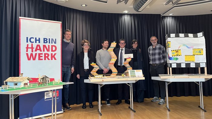 Competenza artigianale e creatività: premiazione del concorso “Abenteuer Handwerk” Gruppo di sei persone dietro tre tavoli con modelli e striscione Ich bin Handwerk