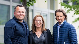 News Drei lächelnde Personen stehen vor einem Gebäude im Freien