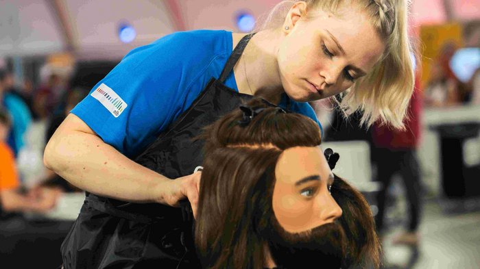 EuroSkills 2023: adesso bisogna concentrarsi! Parrucchiera che acconcia con concentrazione una testa di manichino maschile con capelli lunghi e barba