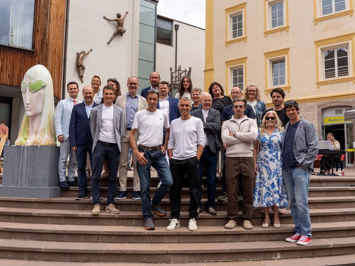 IDEA UNIKA 2025: 6ª edizione della Biennale d’arte nei centri dei paesi della Val Gardena Gruppo di persone posa sulle scale davanti a edifici durante mostra d'arte