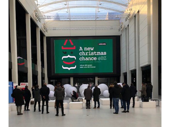 A NEW CHRISTMAS CHANCE: ES GEHT LOS! Menschen stehen vor einer großen Leinwand mit Weihnachtswerbung in einem hellen Raum