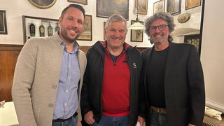 News Drei Männer stehen lächelnd in einem Raum mit Bildern an der Wand