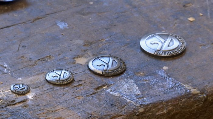 55 Jahre Schutzmarke für das Südtiroler Kunsthandwerk Vier runde Metallmarken unterschiedlicher Größe auf Holzoberfläche